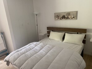 Una cama bien hecha con edredón blanco y almohadas, una lámpara de noche y una obra de arte en la pared.