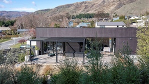 Un edificio moderno con una zona de estar exterior cubierta, rodeado de vegetación y un paisaje montañoso.