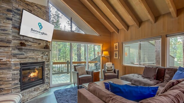 A modern living room with a stone fireplace, wooden ceiling beams, and large windows offering a view of the outdoors. - LEIS06 - Great for Families, Hot Tub, Ping Pong, Bunk Rooms (Sunriver)