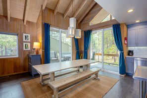 A wooden interior with a large table and bench, a kitchen area, and large windows with curtains.