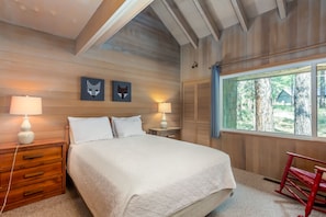 A bedroom with a large bed, two bedside tables with lamps, a wooden dresser, and a window with a view of trees.