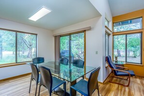 Une pièce moderne avec une table à manger en verre, quatre chaises, un fauteuil bleu et de grandes fenêtres donnant sur la forêt.