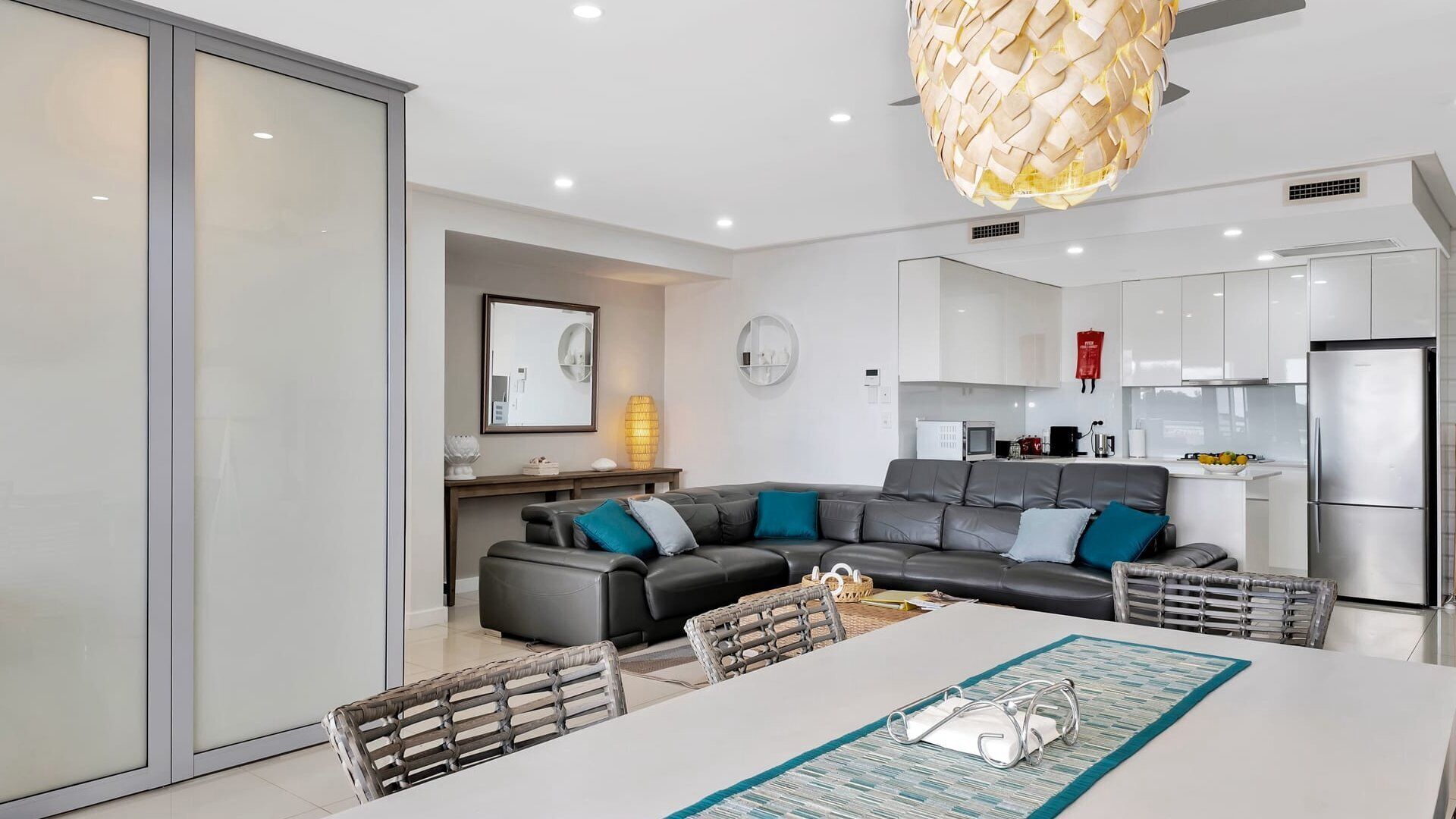 A modern kitchen with stainless steel appliances, a dining area with a white table and wicker chairs, and a gray leather sofa with teal cushions.