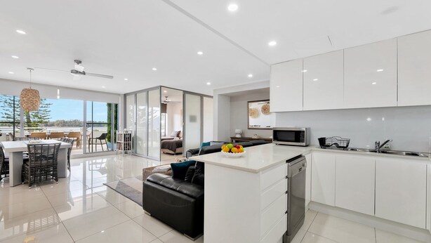 Una cocina moderna con armarios blancos, una isla central con un frutero, y un comedor con un ventanal que da a un balcón.