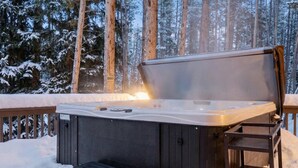 Ein Whirlpool in verschneiter Winterlandschaft mit einer Holzterrasse und hohen Bäumen im Hintergrund.