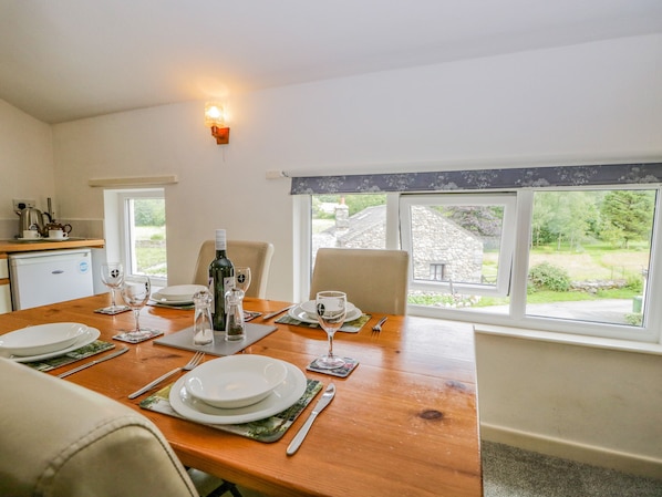 Comedor con mesa de madera dispuesta para dos, una kitchenette pequeña y vista a un edificio de piedra y vegetación al exterior.