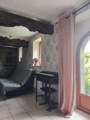 A room with a stone fireplace, a piano, a cushioned chair, and a window with a curtain.