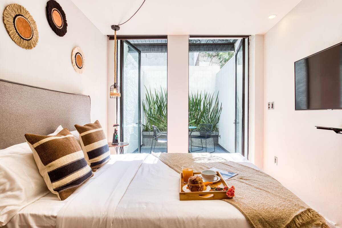 A modern hotel room with a bed, pillows, a tray with breakfast, a TV, and a view of the outdoors.