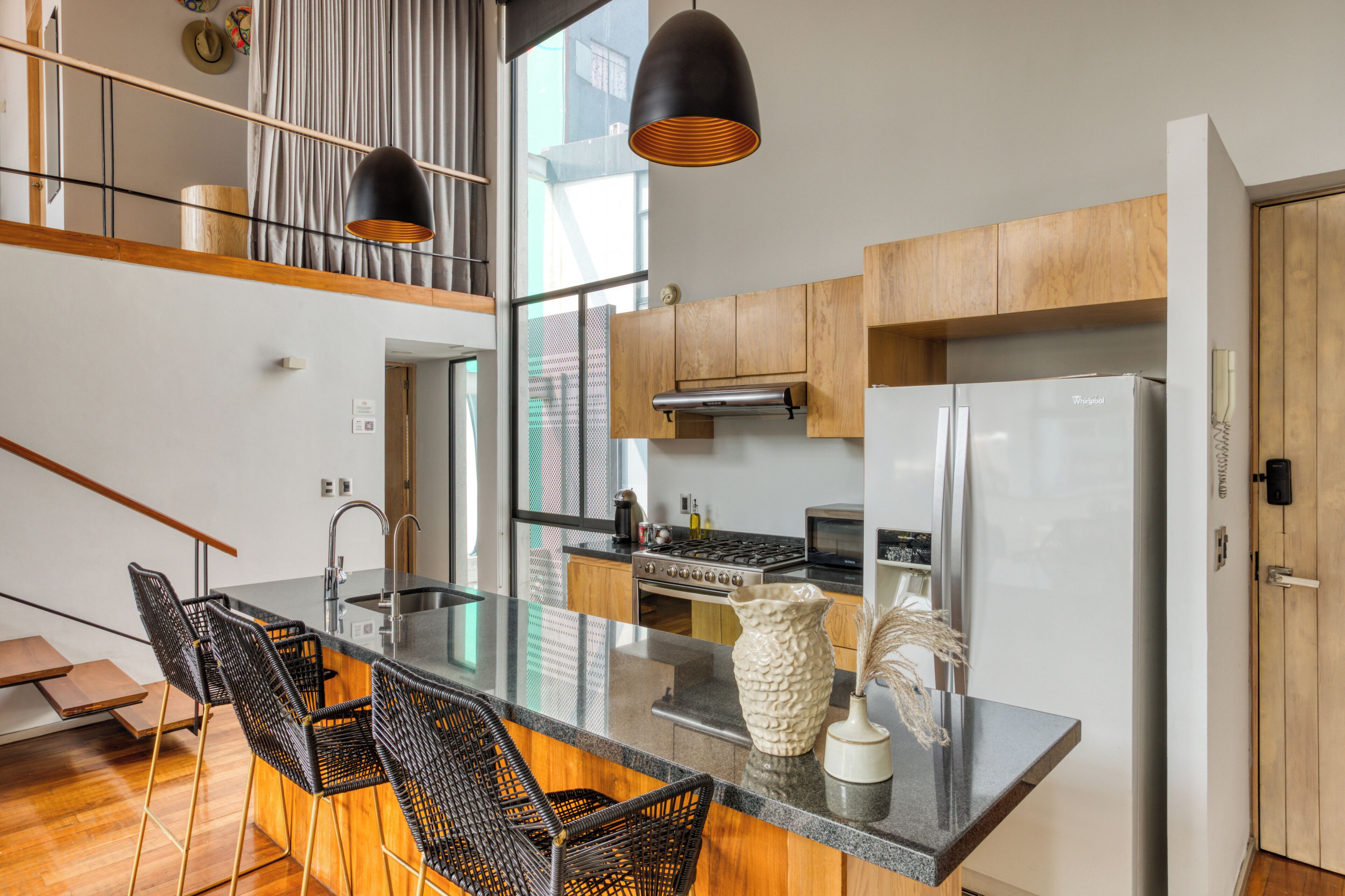 A modern kitchen with a central island, wooden cabinets, stainless steel appliances, and black wicker chairs.