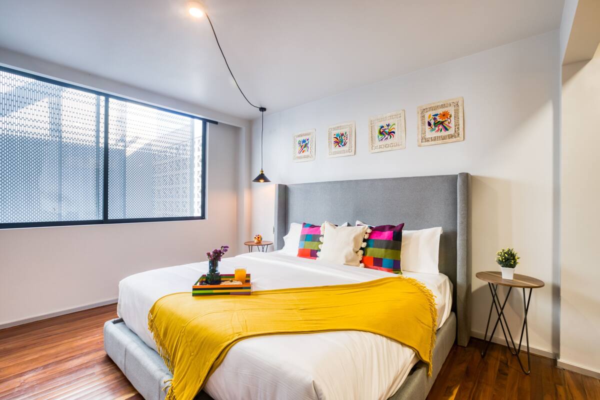 A modern bedroom with a large bed, a small side table, a window with blinds, and wall art.