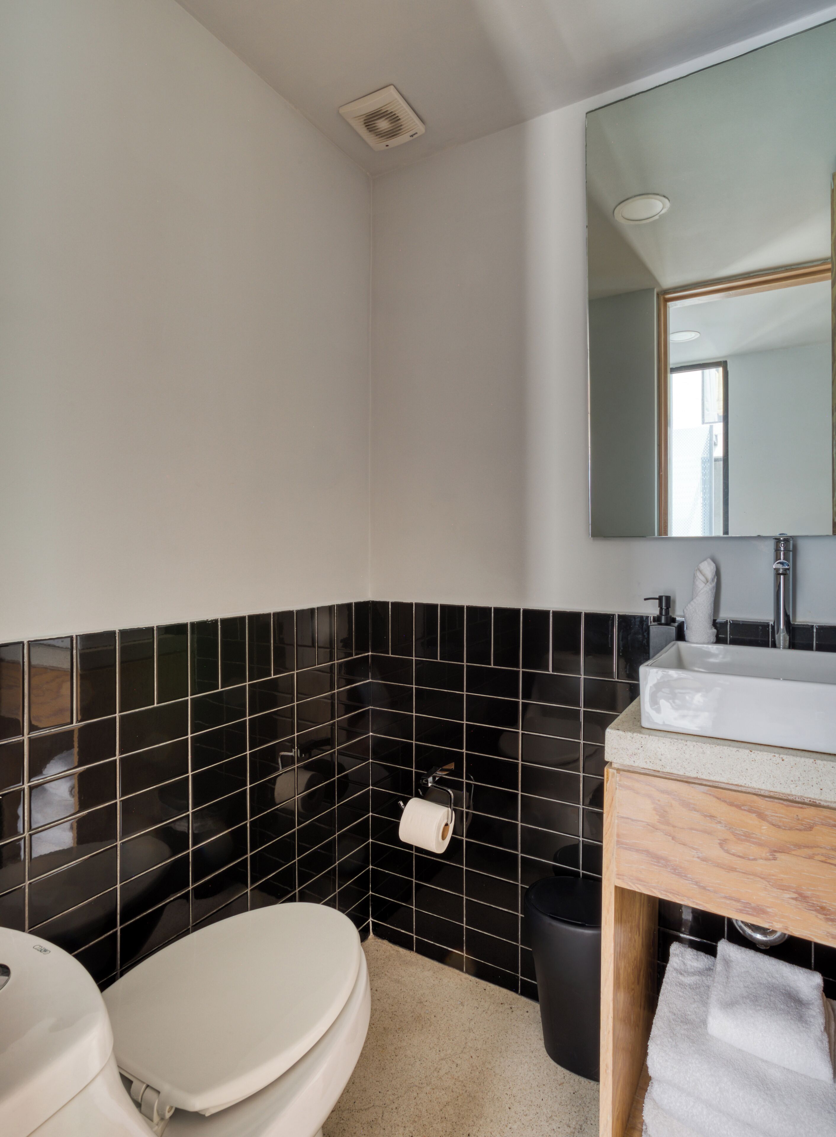 Un baño con inodoro, lavabo, espejo y paredes con azulejos negros.