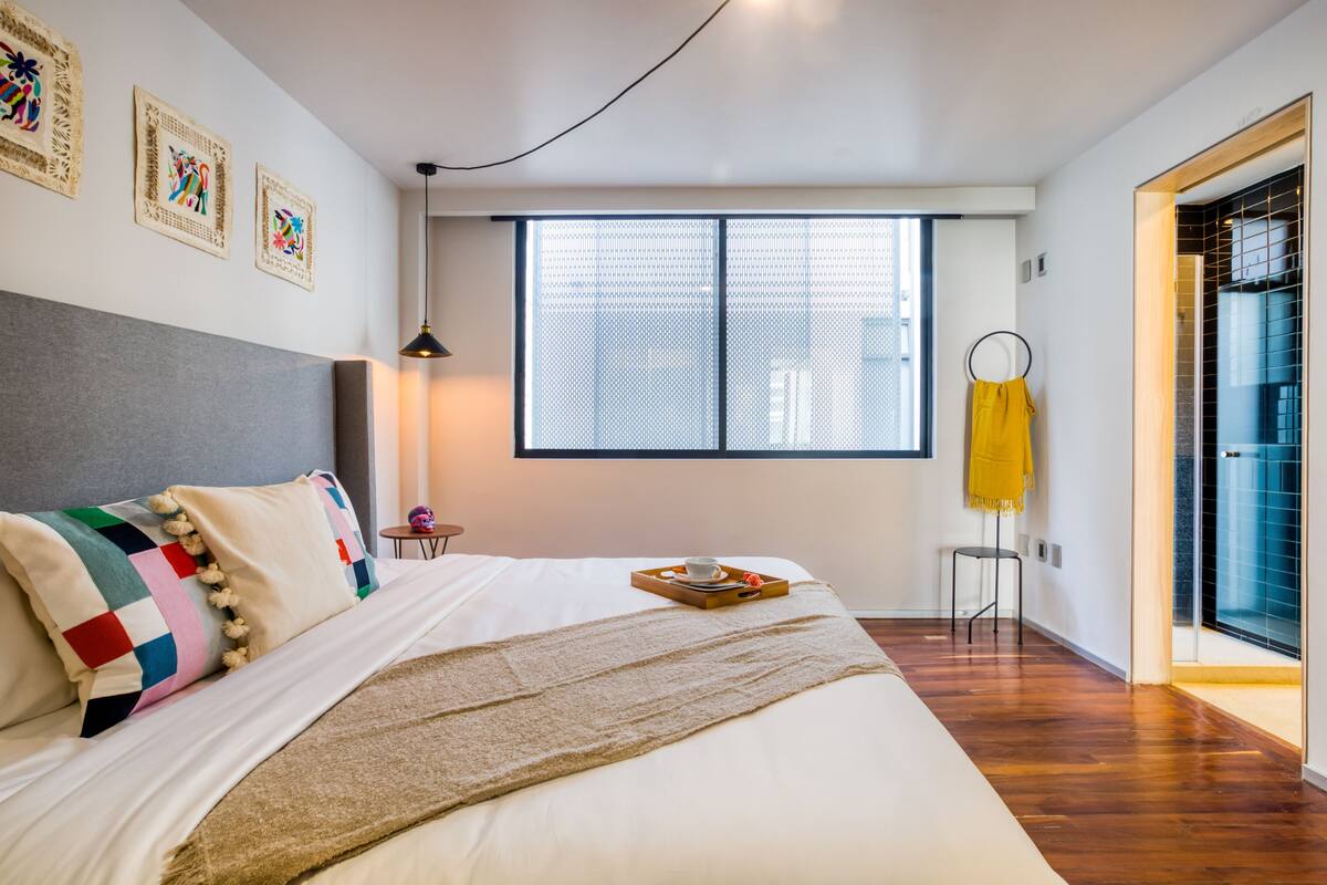 A modern hotel room with a large bed, a window with a view, a small table with a tray, and a yellow towel hanging on the wall.