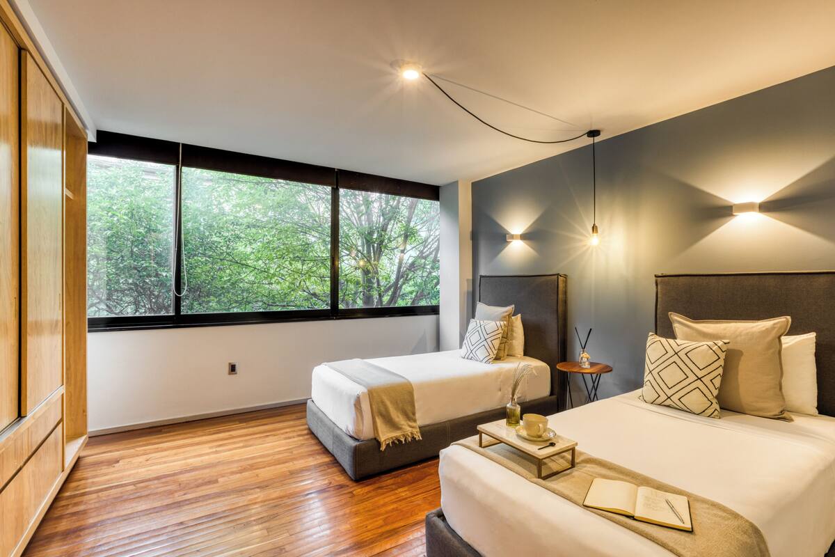 A modern hotel room with two beds, a large window, and a view of trees.