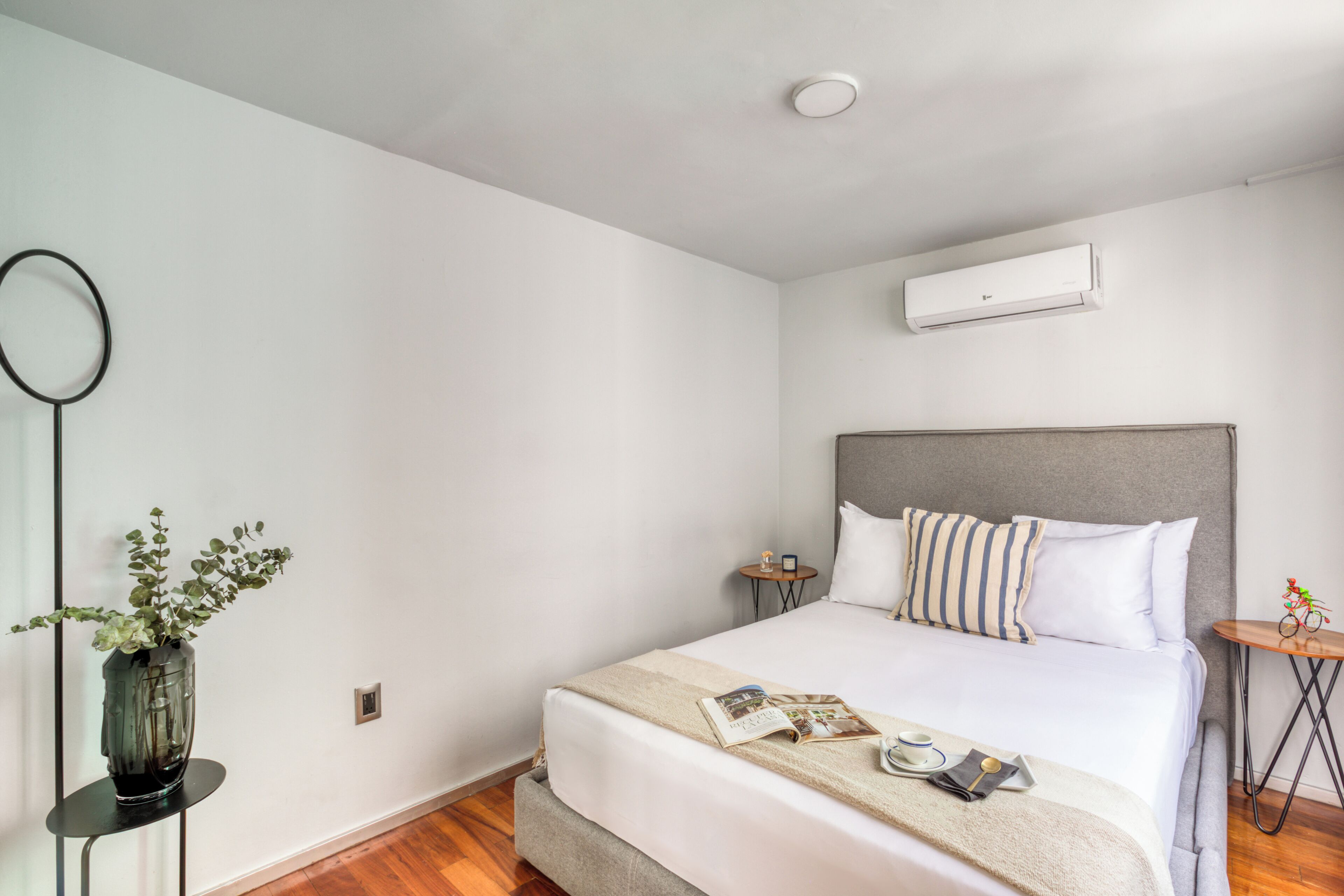 A neatly arranged hotel room with a bed, bedside tables, a mirror, and an air conditioning unit.