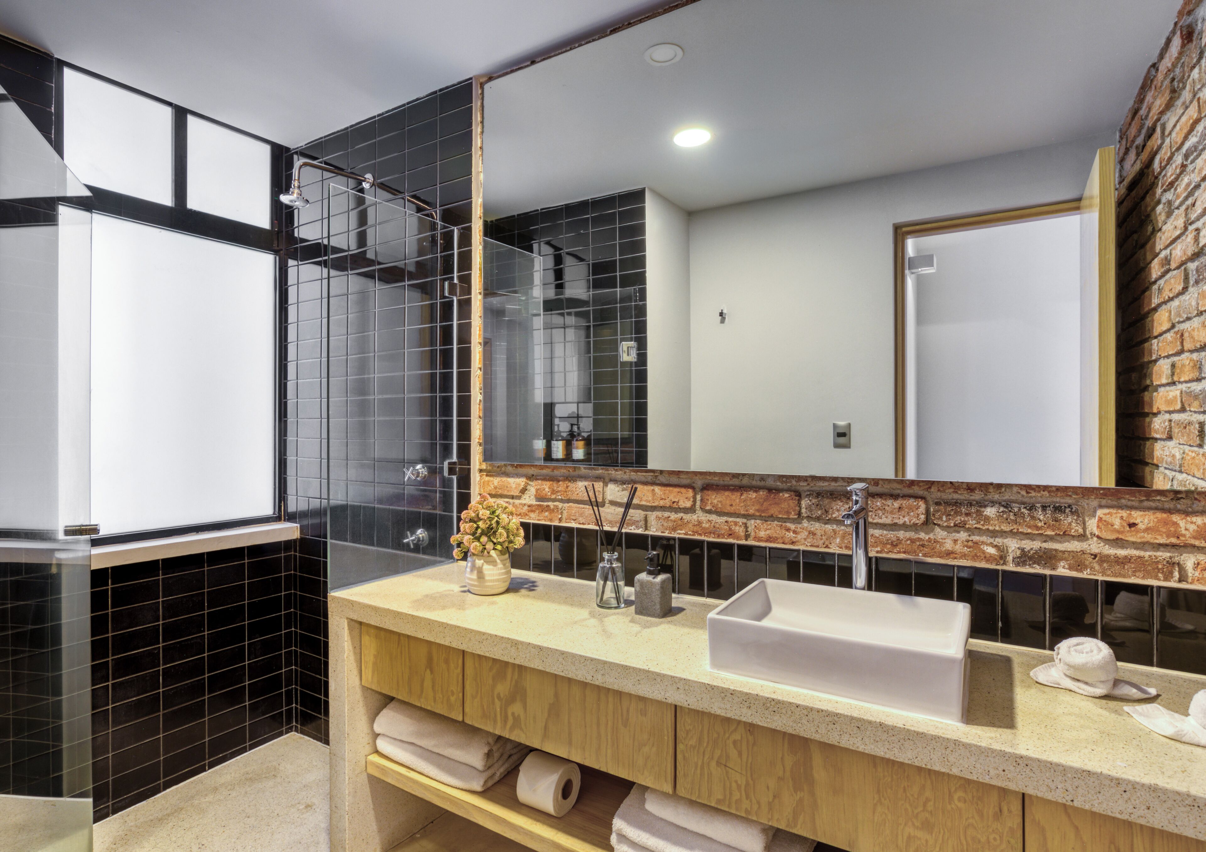 Une salle de bain moderne avec un grand miroir, des murs recouverts de carreaux noirs, un meuble-lavabo en bois et un mur d’accent en briques.