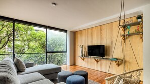 A modern living room with a large window, a grey sofa, ottomans, a hanging chair, and a mounted TV. - ULIV Parque México (Mexico City)