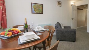 Un comedor con una mesa de madera dispuesta para dos personas, una botella de champán y fruta sobre la mesa. Hay un sofá gris y una puerta que da a otra habitación.