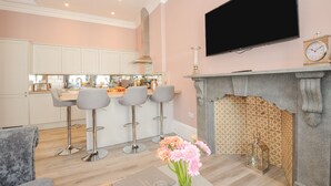A modern kitchen with white cabinets, a central island with bar stools, a flat-screen TV, and a patterned fireplace mantel. - The Stannary (Helston)