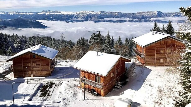Tres cabañas de madera con techos cubiertos de nieve, rodeadas por un paisaje nevado y montañas al fondo.