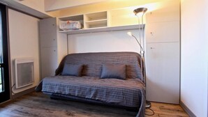 Un dormitorio pequeño con cama, un radiador, una estantería con objetos y una lámpara de pie.
