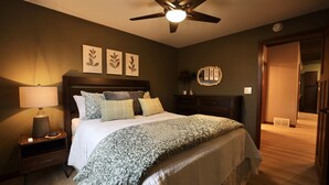 A bedroom with a bed, nightstand, lamp, ceiling fan, and artwork on the wall. - Modern Rustic 2BR Condo in Downtown Minocqua—Walk to Shops, Dining & Lake (Minocqua)