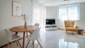Una sala de estar moderna con una mesa de comedor de madera, sillas blancas, un televisor sobre un mueble blanco, un sillón amarillo y un ventanal con persianas.