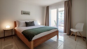 Un dormitorio con una cama de madera, una mesita de noche con lámpara, una silla y vistas al exterior a través de un ventanal amplio.