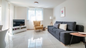 Una sala de estar moderna con un televisor de pantalla plana, un sofá gris, un sillón color beige y una pequeña mesa redonda.
