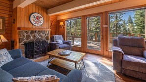 Interior acogedor de una cabaña con chimenea de piedra, paredes de madera y ventanas amplias que dan al bosque.