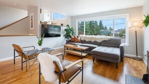 A modern living room with a grey sofa, a wooden coffee table, and a brown leather chair. A staircase is visible to the left, and a large window provides a view of the outdoors. - Perch - Gathering Spot: Spacious 5BR home in Shoreline (Shoreline)