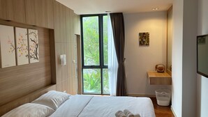 Habitación de hotel moderna con una cama grande, cabecera de madera, ventana con cortinas, un estante pequeño con un objeto decorativo y un cesto de ropa blanca.