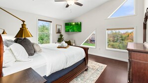 Un dormitorio con una cama grande, un ventilador de techo, un televisor y dos ventanas con vistas a árboles.