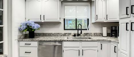 Una cocina moderna con armarios blancos, lavavajillas de acero inoxidable, encimera de granito y ventana con vistas a zonas verdes.