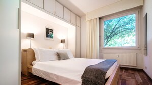 A modern bedroom with a wooden bed, white linens, a small bedside table, two wall-mounted lamps, and a window with a view of trees. - Cadro Garden (Lugano)