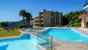 A swimming pool with a glass barrier, surrounded by a pebble-tiled deck. In the background, there is a multi-story building with balconies and a lush green landscape. - Cadro Garden (Lugano)