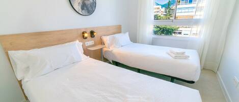 A hotel room with two beds, a wooden headboard, a wall-mounted light fixture, and a window with a view of buildings and greenery.