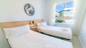 A hotel room with two beds, a wooden headboard, a wall-mounted light fixture, and a window with a view of buildings and greenery. - PREMIUM Apartment with 3 bedrooms. Capacity for 6 people (Marbella)