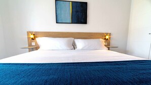 Un lit bien fait, avec du linge de lit blanc et une couverture bleue, une tête de lit en bois et deux lampes de chevet avec des abat-jour jaunes.