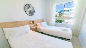 A hotel room with two beds, a wooden headboard, a wall-mounted light fixture, and a window with a view of buildings and greenery. - Deluxe 2 Bedroom Apartment. Capacity for 4 people (Marbella)