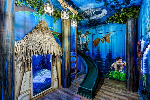 A room with a curved green slide, a straw-roofed bed, and a mural of a forest scene.