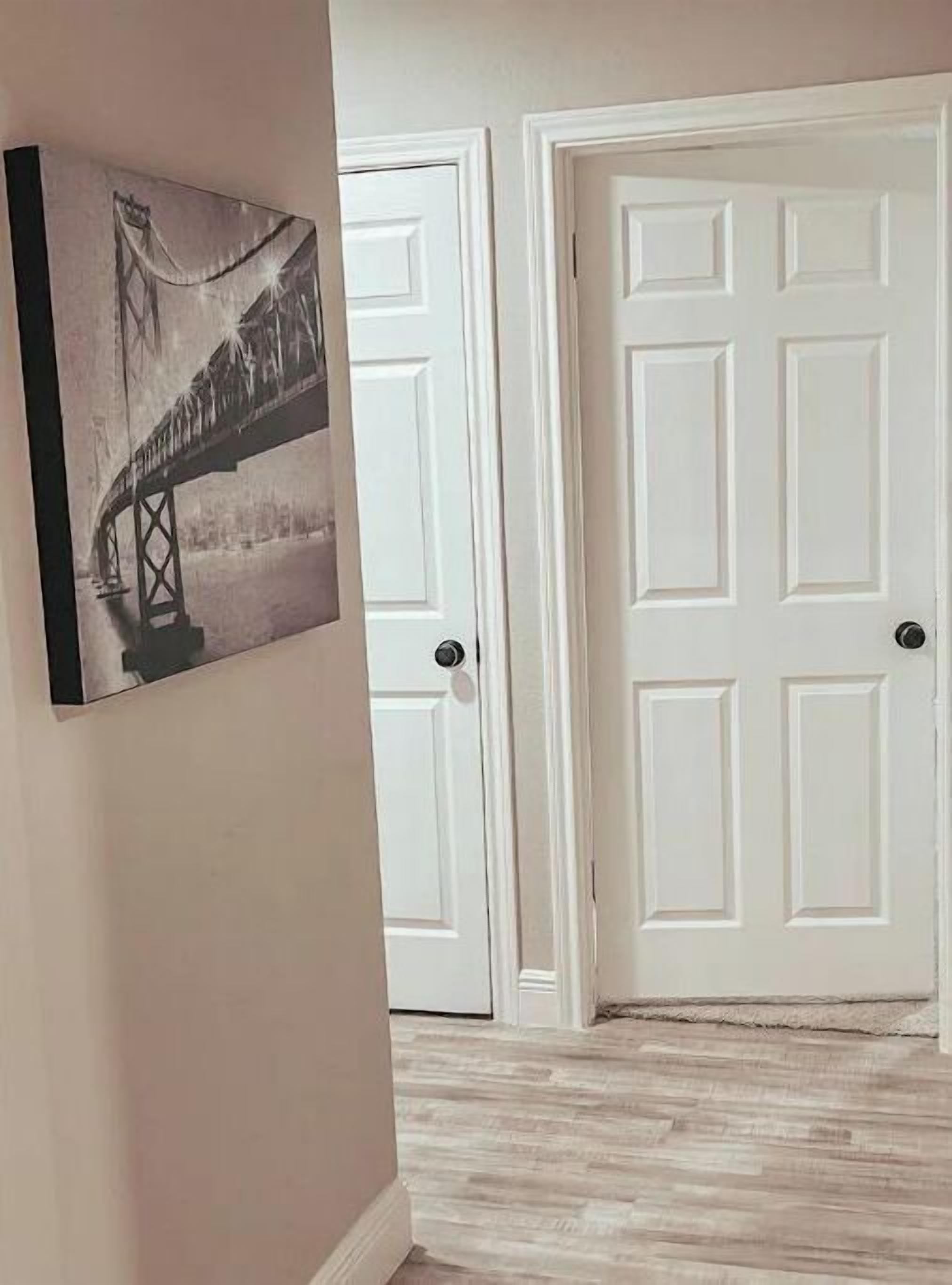 A hallway with a framed black and white photograph of a bridge, white doors, and a light-colored wooden floor.