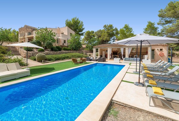 A swimming pool with lounge chairs and umbrellas, surrounded by a landscaped garden and a building with a balcony. - Villa ES PINS in PORTOCOLOM (Portocolom)