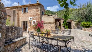 A stone building with a rustic exterior, a small patio with metal chairs and a table, and a garden with pink flowers. - AINA CAN PONTICO Charming villa in Pollença (Pollença)