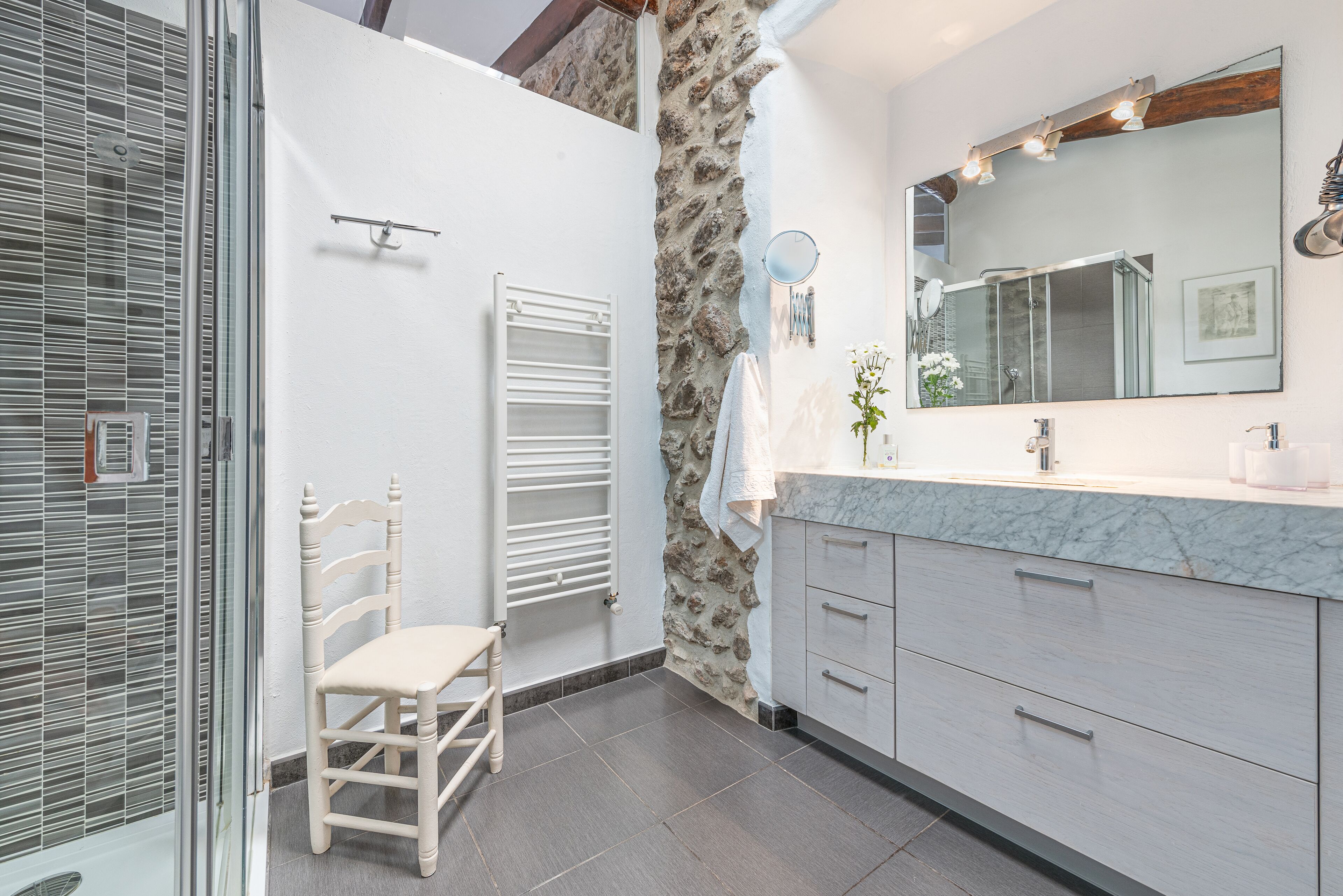 A bathroom with a stone wall, a white chair, a large mirror, and modern cabinetry.
