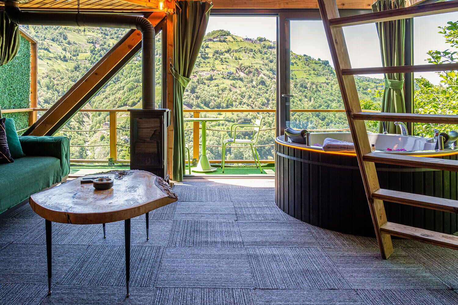 A room with a view of a green hillside, a wooden table, a green sofa, and a wooden ladder.