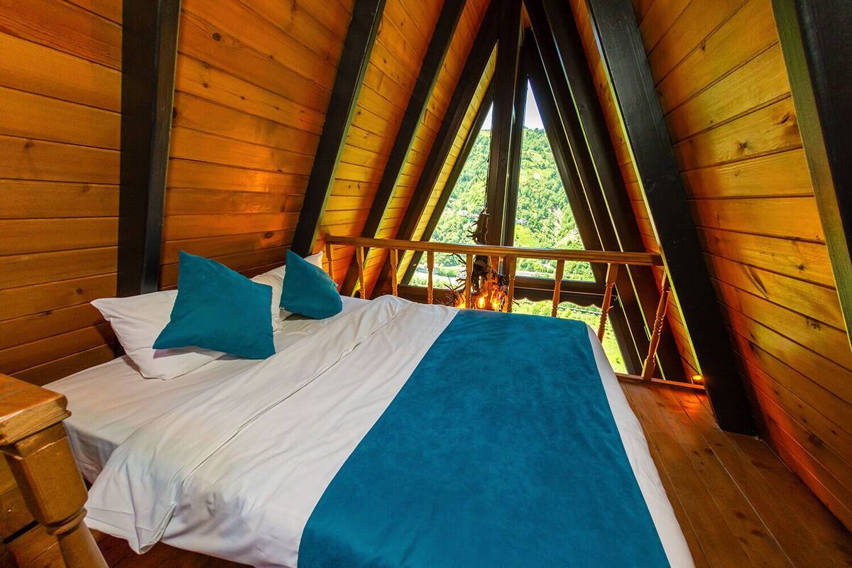 A wooden cabin room with a bed, pillows, and a view of greenery through a window.