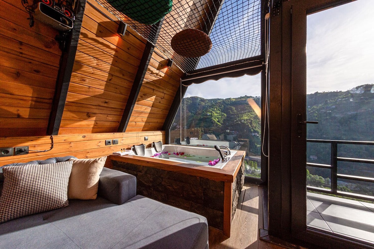 A wooden cabin with a stone bench, a bathtub with flowers, and a view of a mountainous landscape.