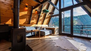 A cozy cabin with a stone fireplace, a sofa, and a dining table. Large windows offer a view of the surrounding landscape.