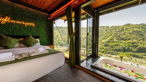 A bedroom with a bed, pillows, a view of a green landscape through large windows, and a neon sign on the wall.