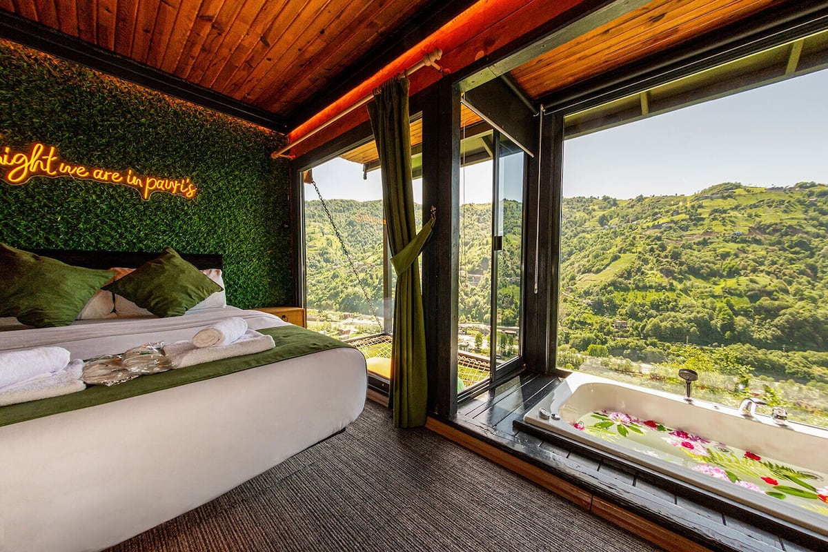 A bedroom with a bed, pillows, a view of a green landscape through large windows, and a neon sign on the wall.